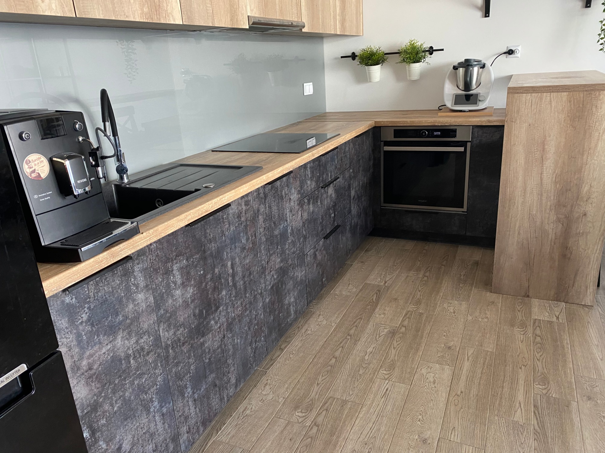 Nowoczesna kuchnia z ciemnymi, teksturowanymi frontami szafek, drewnianym blatem i szarym panelem nad blatem. Na blacie widoczny ekspres do kawy, zlewozmywak, płyta indukcyjna oraz robot kuchenny.