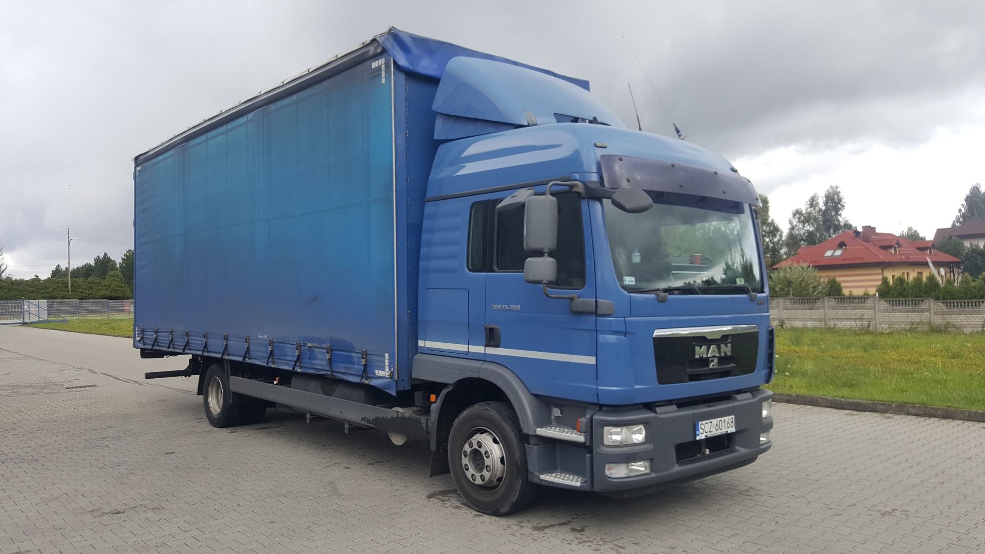 Niebieski samochód ciężarowy MAN z plandeką, zaparkowany na kostce brukowej, z widocznym numerem rejestracyjnym SCZ 60168 i budynkami mieszkalnymi w tle.