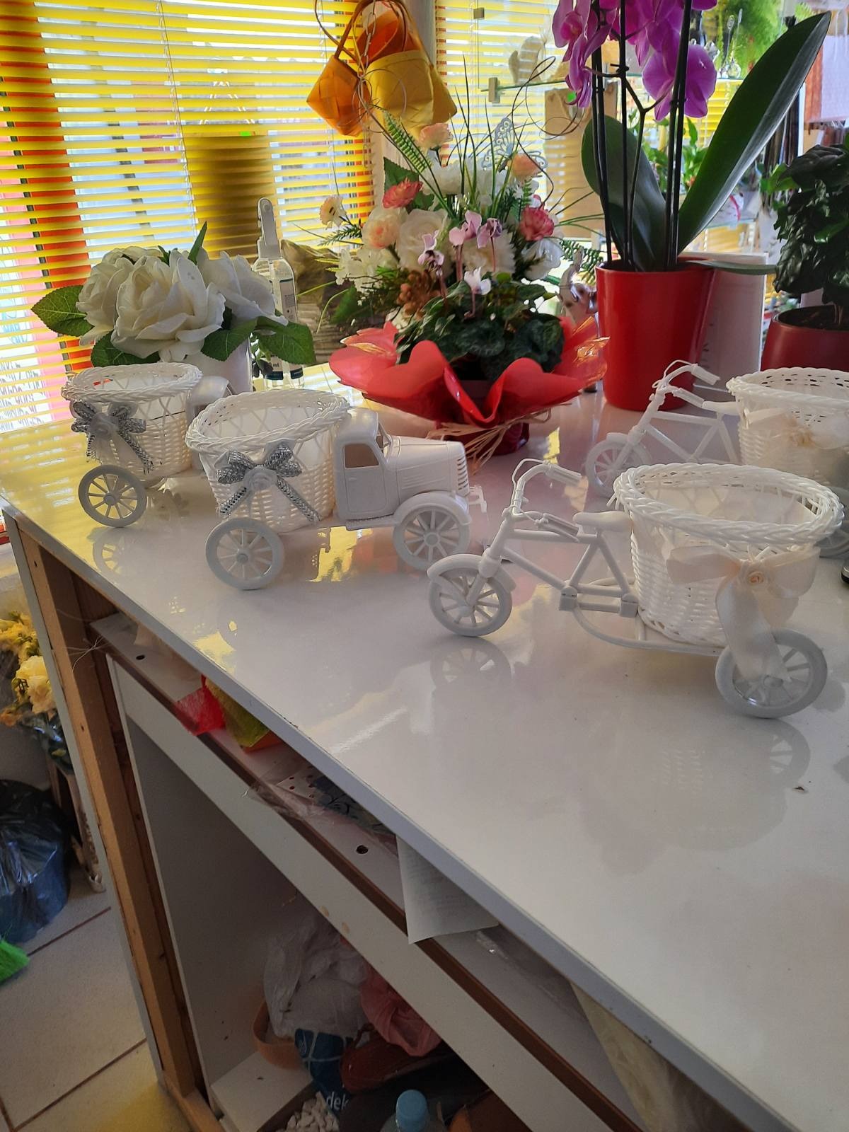 Białe, dekoracyjne wózki i rowerki z wiklinowymi koszyczkami, potencjalnie jako doniczki, ustawione na białym blacie, z kwiatami w tle.