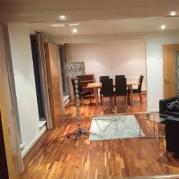 Salon z parkietem, widok na jadalnię z drewnianym stołem i czarnymi krzesłami, lampa podłogowa z wzorzystym abażurem i szklany stolik kawowy na geometrycznym dywanie.