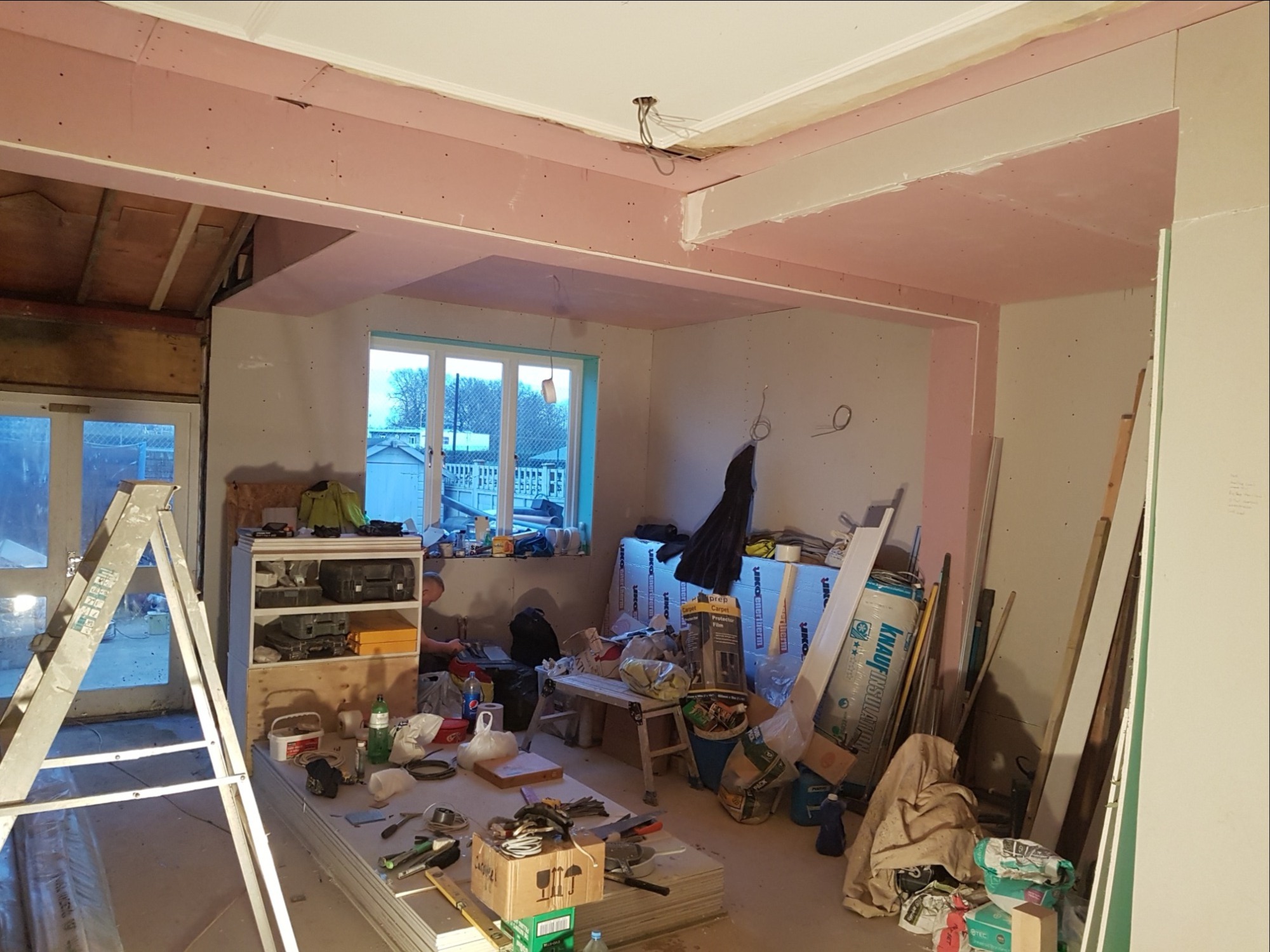 Wnętrze w trakcie prac wykończeniowych: widoczne płyty kartonowo-gipsowe na suficie i ścianach, drabina, narzędzia, materiały budowlane i osoba pracująca w tle.