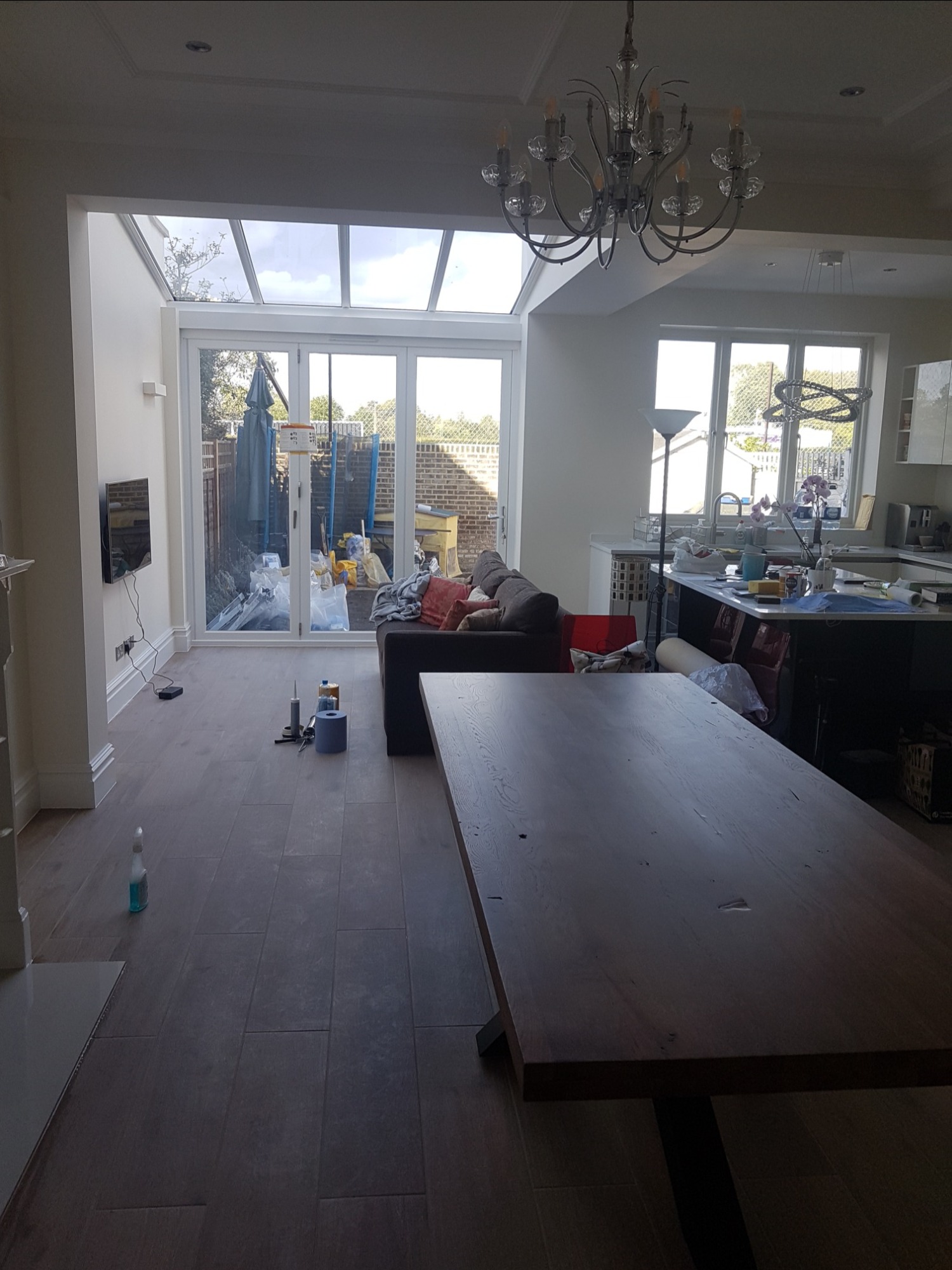 Wnętrze domu w trakcie prac wykończeniowych: widoczny duży stół na pierwszym planie, salon z sofą, telewizorem i przeszklonym wyjściem na ogród, oraz częściowo widoczna kuchnia z wyspą.