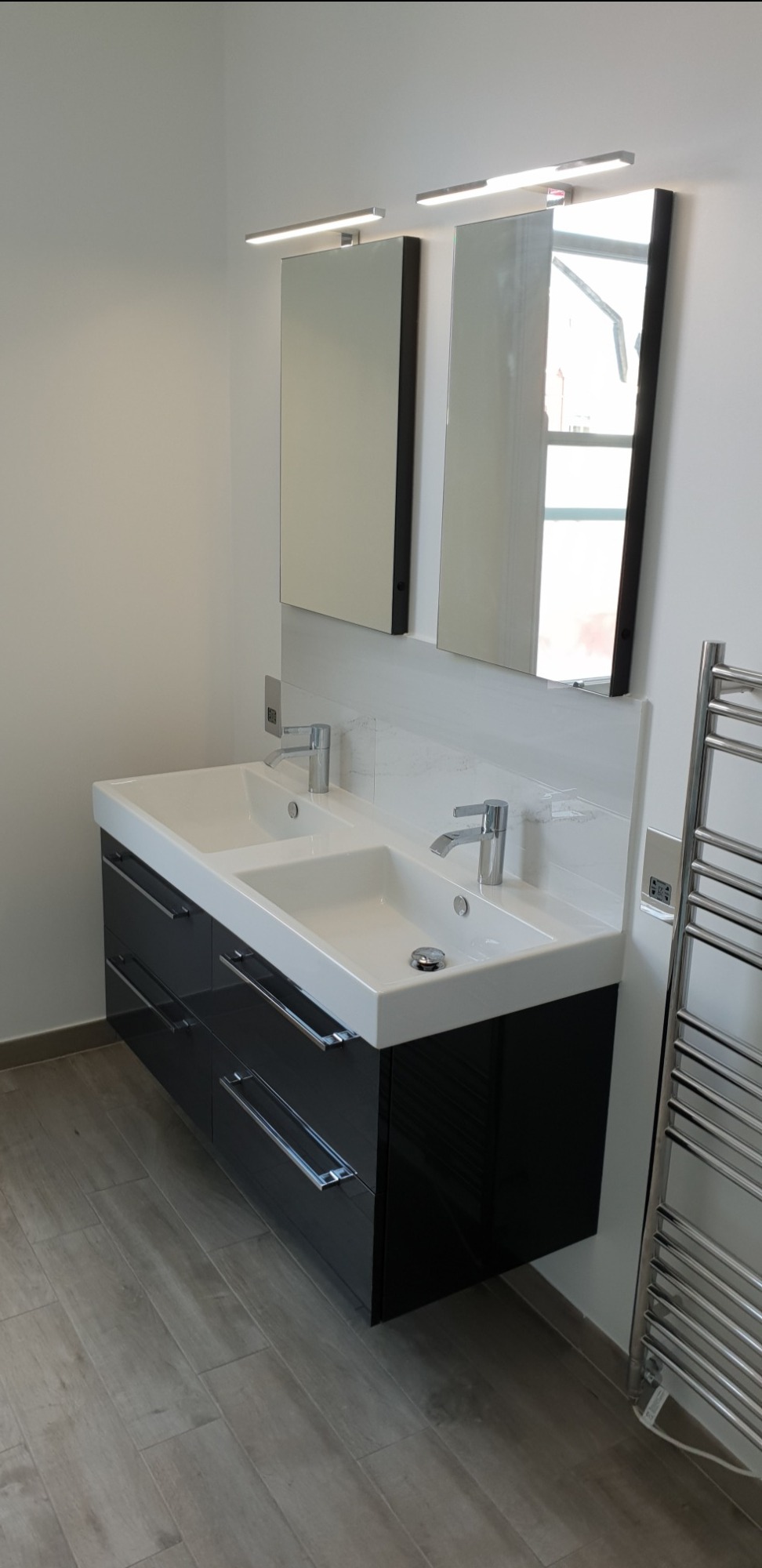 Nowoczesna łazienka z podwójną umywalką nablatową, czarnymi szafkami z chromowanymi uchwytami, dwoma prostokątnymi lustrami z oświetleniem LED i grzejnikiem drabinkowym.