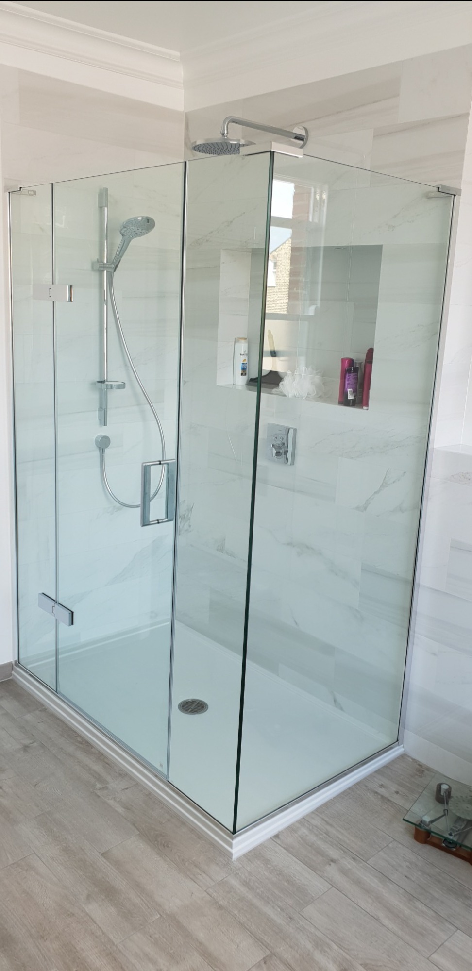 Nowoczesna kabina prysznicowa z przezroczystego szkła, chromowane wykończenia, białe płytki imitujące marmur oraz wnęka na kosmetyki w ścianie.