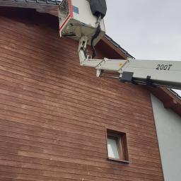 PHU Katarzyna Halka - Praca na wysokościach przy drewnianej elewacji domu jednorodzinnego z użyciem podnośnika koszowego 200T, widoczny pracownik w koszu.