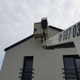 Biały podnośnik koszowy z numerem telefonu na ramieniu, ustawiony przy ścianie nowoczesnego domu z ciemnymi oknami i szarym niebem w tle.