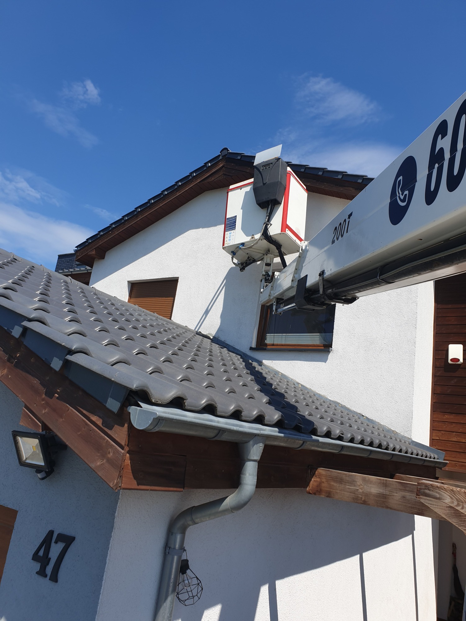 Biały dom z szarą dachówką i numerem 47, z bliska widoczny podnośnik koszowy z wysięgnikiem sięgającym okna, błękitne niebo w tle.