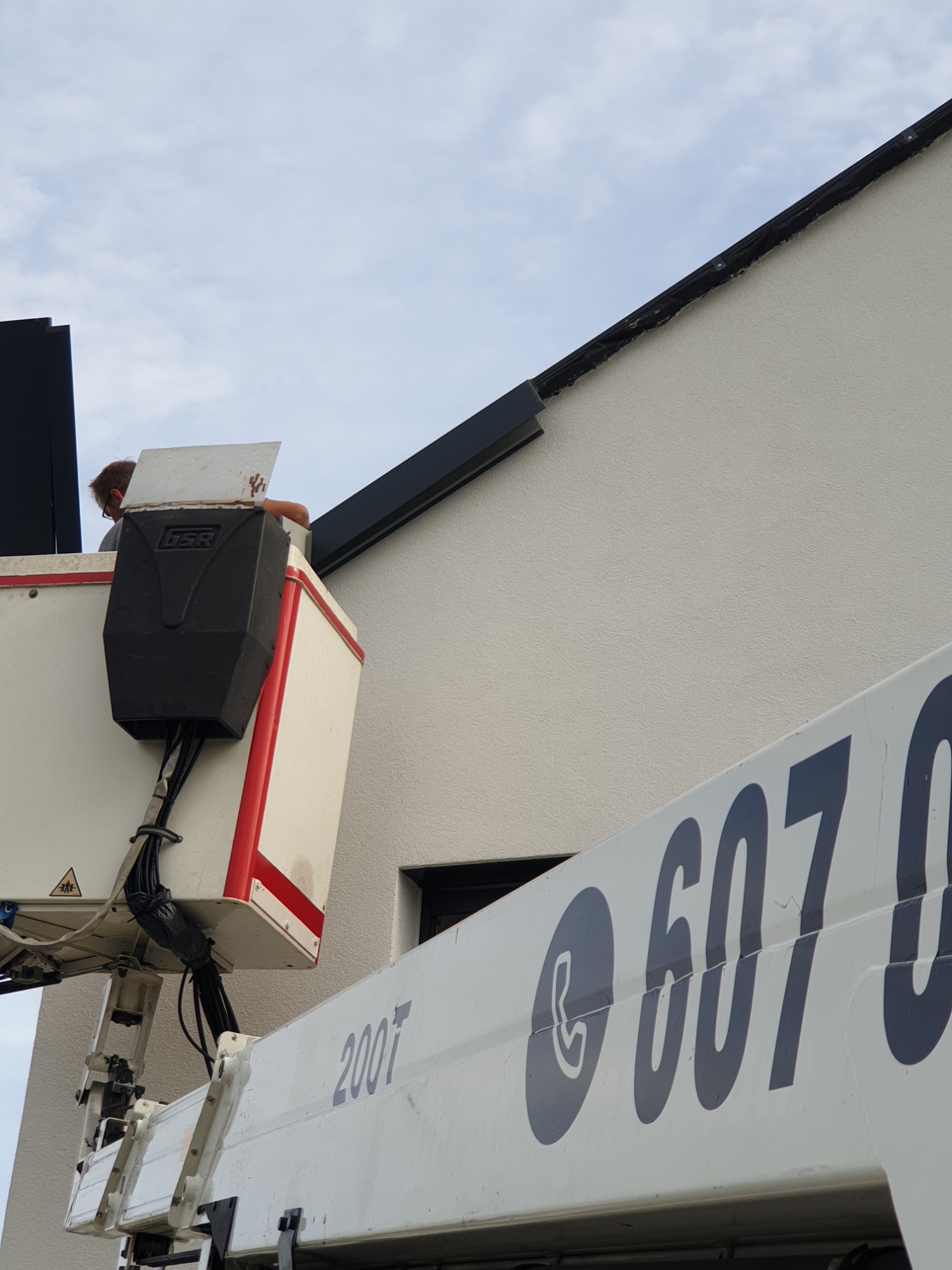 Pracownik w podnośniku koszowym, montujący obróbkę blacharską dachu na elewacji budynku, widoczny numer telefonu na maszynie.