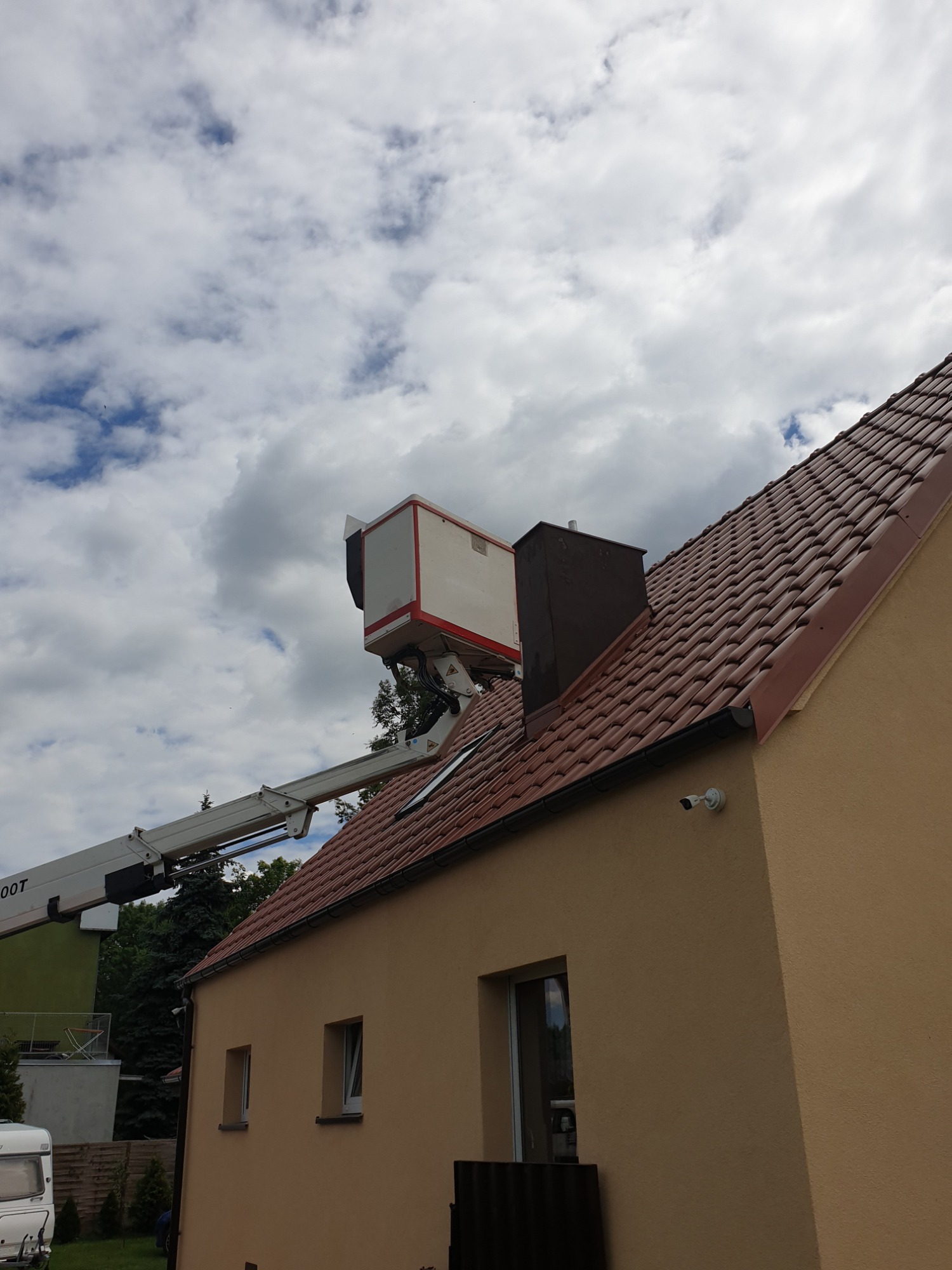 Podnośnik koszowy z biało-czerwoną platformą roboczą ustawiony przy brązowym dachu domu jednorodzinnego, widoczny fragment komina i kamera monitoringu na elewacji.