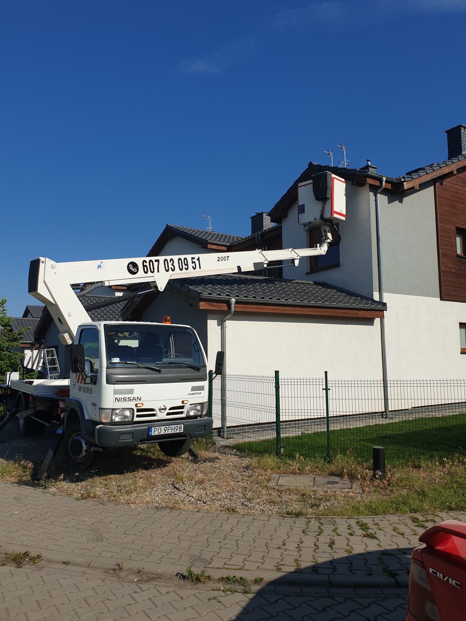 Biały samochód z wysięgnikiem z numerem telefonu na tle jasnego domu jednorodzinnego z ciemną dachówką i zielonym ogrodzeniem.