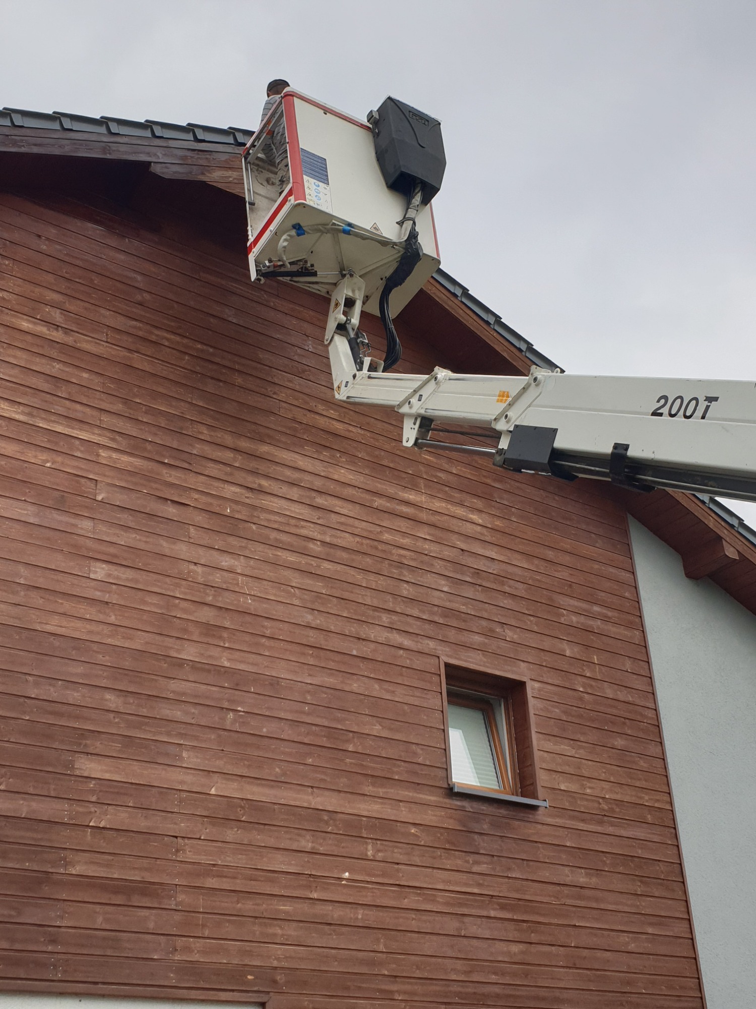 Praca na wysokościach przy drewnianej elewacji domu jednorodzinnego z użyciem podnośnika koszowego 200T, widoczny pracownik w koszu.