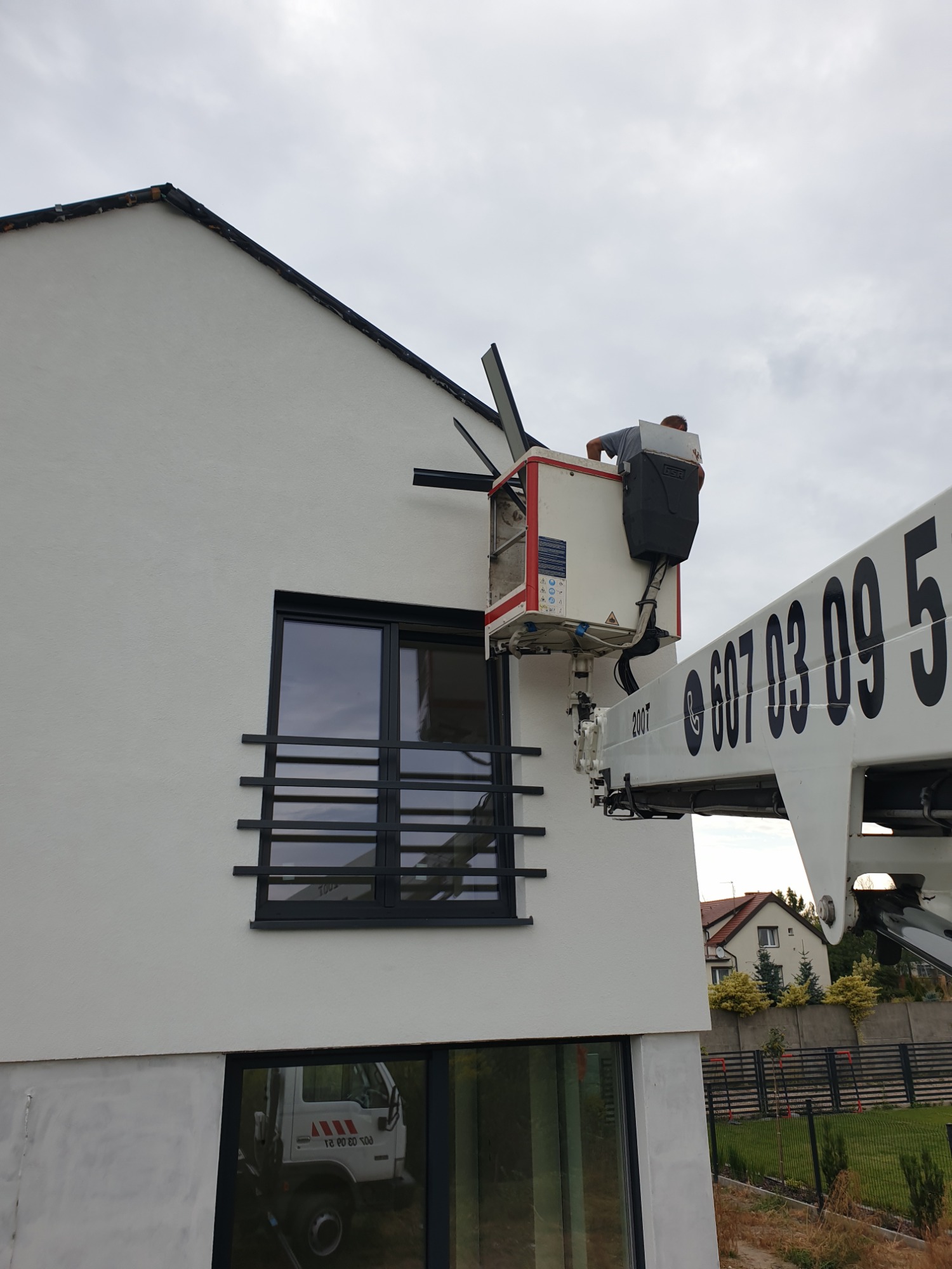 Pracownik w koszu podnośnika przy elewacji dwukondygnacyjnego domu z czarną stolarką okienną, widoczny numer telefonu na ramieniu podnośnika.