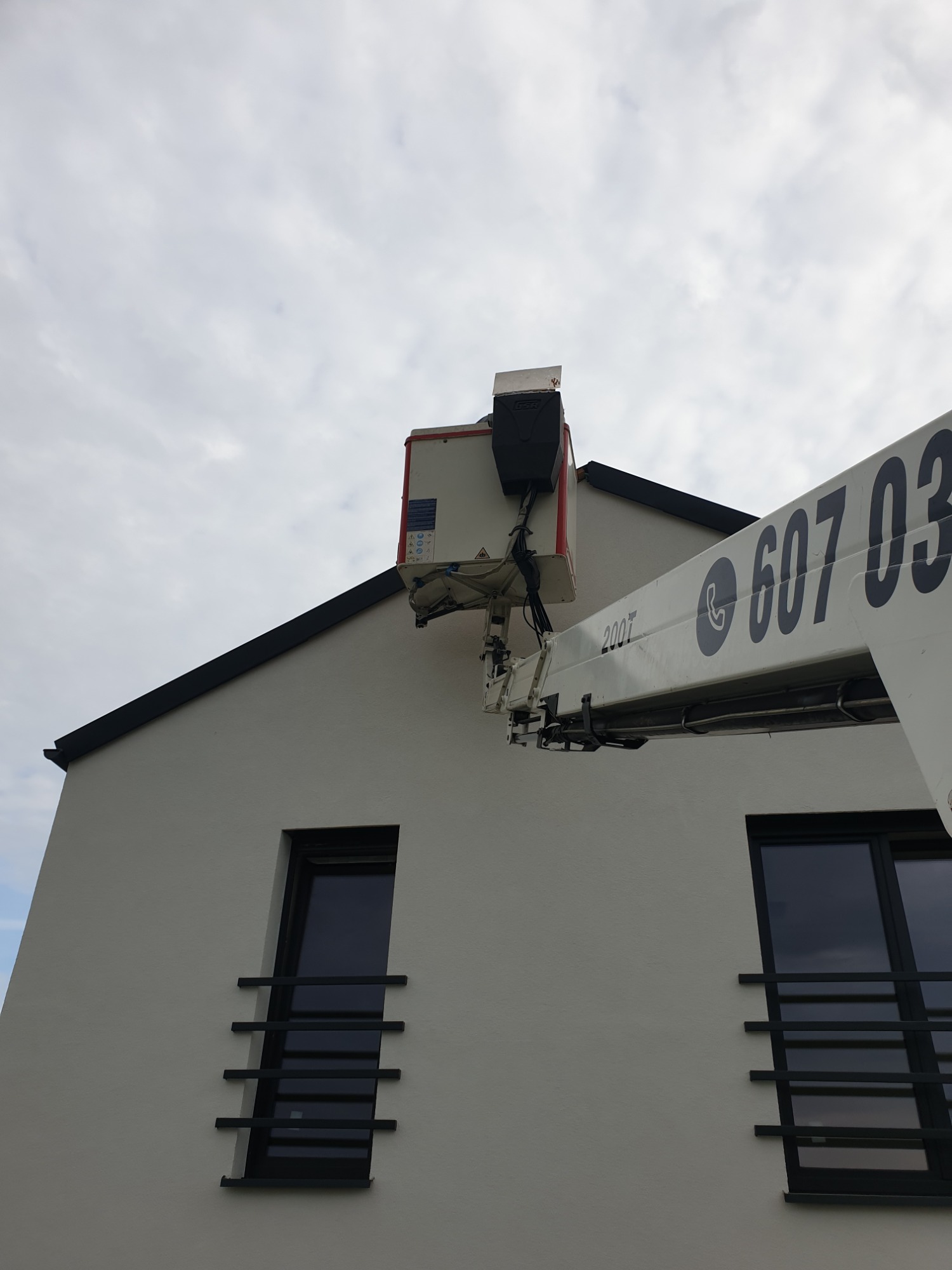 Biały podnośnik koszowy z numerem telefonu na ramieniu, ustawiony przy ścianie nowoczesnego domu z ciemnymi oknami i szarym niebem w tle.