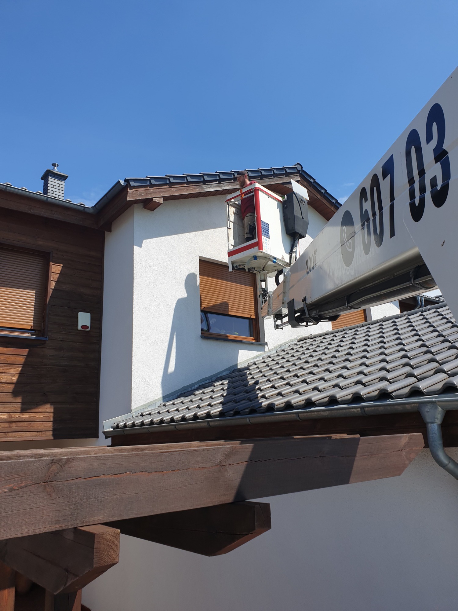 Pracownik w koszu platformy roboczej na wysokości, przy oknie z żaluzjami zewnętrznymi, cień osoby na ścianie budynku.