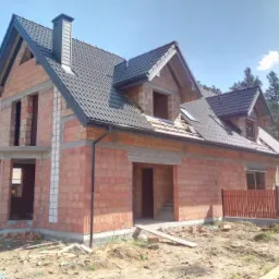 Surowy dom z czerwonej cegły z ciemną dachówką, widoczne niezamknięte otwory okienne i drzwiowe, częściowo ukończony dach z kominem, na pierwszym planie ziemia i drewniane elementy budowlane.