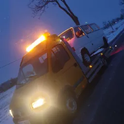 Żółta laweta Renault z włączonymi światłami i kogutem holuje białego Volkswagena Caravelle po ośnieżonej drodze o zmierzchu.