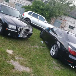 Trzy samochody osobowe (Chrysler, BMW, Porsche Cayenne) zaparkowane na trawiastym terenie przed budynkiem z cegły z widocznymi garażami.