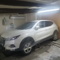 Biały samochód owinięty folią ochronną w garażu z widocznymi narzędziami i elementami wyposażenia warsztatowego.