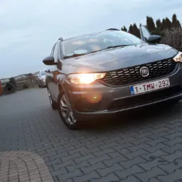 Szary samochód marki Fiat Tipo kombi z otwartymi drzwiami kierowcy, zaparkowany na brukowanej nawierzchni przed domem z brązowymi elementami i tujami w tle, widoczne tablice rejestracyjne belgijskie.