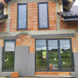 Wstawione okna w surowym budynku z cegły, widoczne ocieplenie styropianem, brak tynków zewnętrznych.