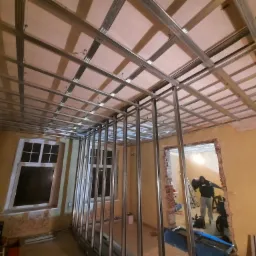 Stalowa konstrukcja sufitu podwieszanego i ścian działowych na tle starego, częściowo otynkowanego pomieszczenia z widoczną cegłą i oknem, w tle pracownik z drabiną.