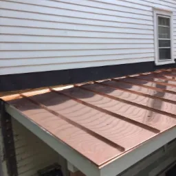 Nowy, błyszczący, miedziany dach panelowy na werandzie z białymi deskami okapowymi, widoczny fragment ściany domu z białym sidingiem i oknem.