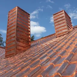 Miedziany dach z dwoma kominami obłożonymi miedzianą blachą, widoczne łączenia blachy na kominach, niebo z chmurami w tle.
