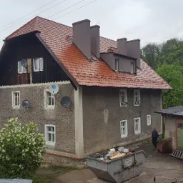 Dwupiętrowy budynek z nowym, czerwonym dachem i trzema kominami, widoczny z boku, z kontenerem na odpady budowlane na pierwszym planie i zielonymi drzewami w tle.