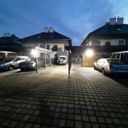 Oświetlony podjazd z kostki brukowej prowadzący do zadaszonych miejsc parkingowych przy budynkach mieszkalnych, widok nocny.