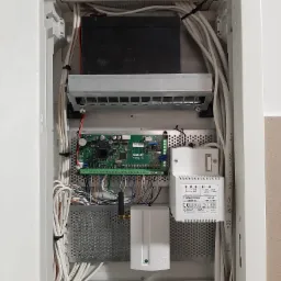 Otwarta szafka z zamontowaną centralą alarmową Satel Versa 10, transformatorem Pulsar AWT150, akumulatorem i okablowaniem wewnątrz białej obudowy.