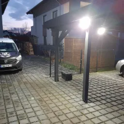 Podjazd do domu z oświetloną wiatą, samochodem dostawczym firmy z logo na masce i skrzynką narzędziową obok słupa wiaty, oświetlenie włączone wieczorem.