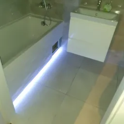 Nowoczesna łazienka z podświetlaną wanną, szarymi płytkami i białą szafką umywalkową z chromowaną baterią i dozownikiem mydła w płynie.