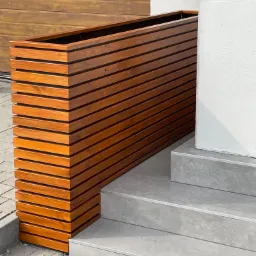 Drewniana donica z poziomych desek, ustawiona przy schodach wejściowych do domu, z widoczną fakturą białej ściany i brązowej bramy garażowej w tle.