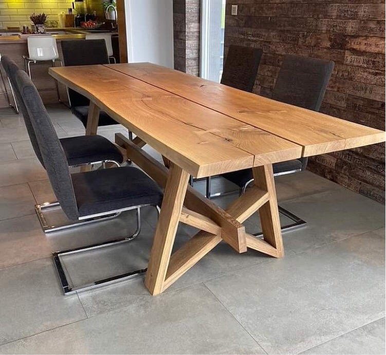 Stół jadalny z jasnego drewna z charakterystycznym krzyżowym stelażem, otoczony krzesłami z szarej tapicerki i chromowanymi nogami, na tle ściany z cegieł i fragmentu kuchni.