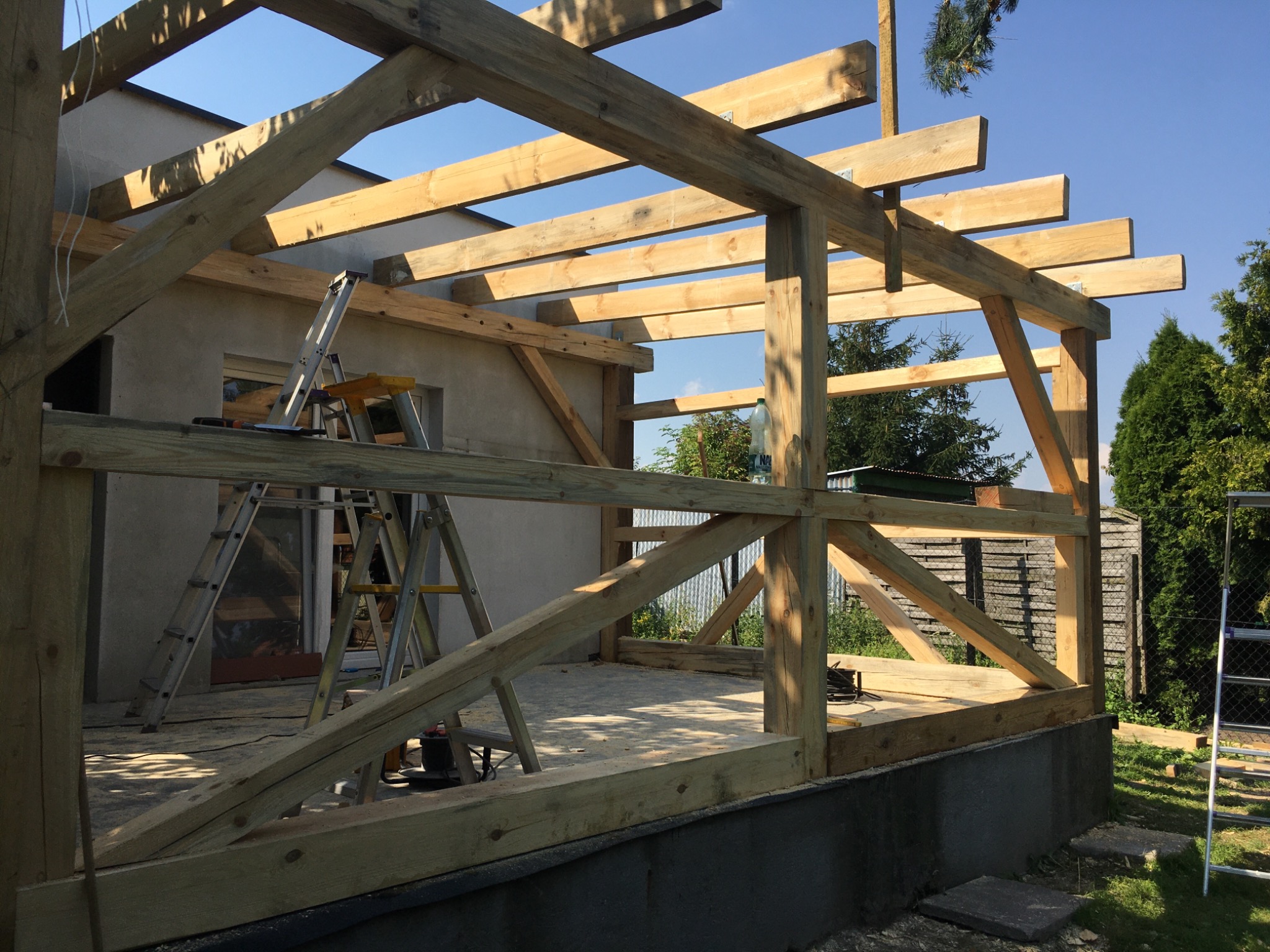Konstrukcja drewniana altany w trakcie budowy, widoczne elementy szkieletu, drabina, narzędzia i betonowa podstawa.