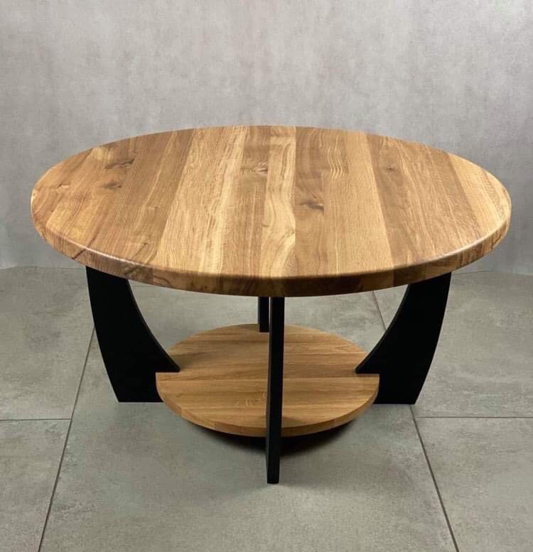 Okrągły stolik kawowy z blatem z jasnego drewna i czarnymi, zakrzywionymi nogami oraz dodatkową półką, na tle szarej ściany i podłogi z płytek.