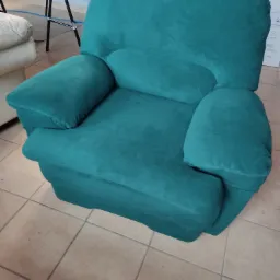 Odnowiony fotel w turkusowym obiciu welurowym, z zaokrąglonymi podłokietnikami, stojący na podłodze wyłożonej jasnymi płytkami ceramicznymi.