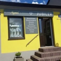 Fasada budynku jubilera z ż&oacute;łtą elewacją, brązowymi ramami okiennymi i schodami wejściowymi. Nad oknem szyld z napisem 'JUBILER' i ofertą usług. Małe drzewko w donicy przy schodach.