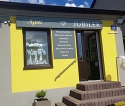 Fasada budynku jubilera z żółtą elewacją, brązowymi ramami okiennymi i schodami wejściowymi. Nad oknem szyld z napisem 'JUBILER' i ofertą usług. Małe drzewko w donicy przy schodach.