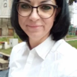 Portret kobiety w okularach i białej koszuli na tle częściowo zasłoniętej tablicy budowlanej i zieleni.