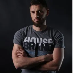 Mężczyzna z brodą, w szarej koszulce z nadrukiem 'House Brand', stoi z założonymi rękami na tle ciemnego studia.