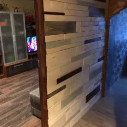 Drewniana ściana działowa z poziomych desek o zróżnicowanej kolorystyce, oddzielająca salon od korytarza z ceglaną ścianą, widoczny fragment brązowej ramy drzwiowej.