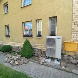 Zewnętrzna jednostka klimatyzacji Panasonic zainstalowana przy ścianie domu z żółtą elewacją, otoczona dekoracyjnym kamiennym obrzeżem i zielonymi krzewami.