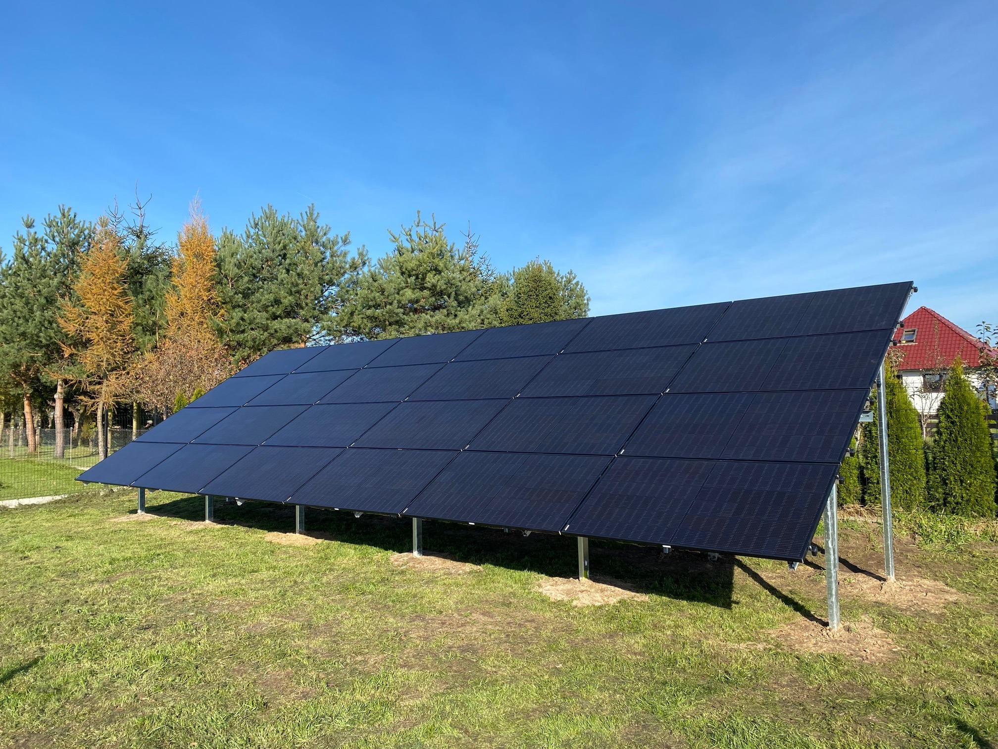 Czarny panel słoneczny zamontowany na metalowych nogach na trawniku, z widocznymi drzewami i fragmentem domu w tle, w słoneczny dzień.