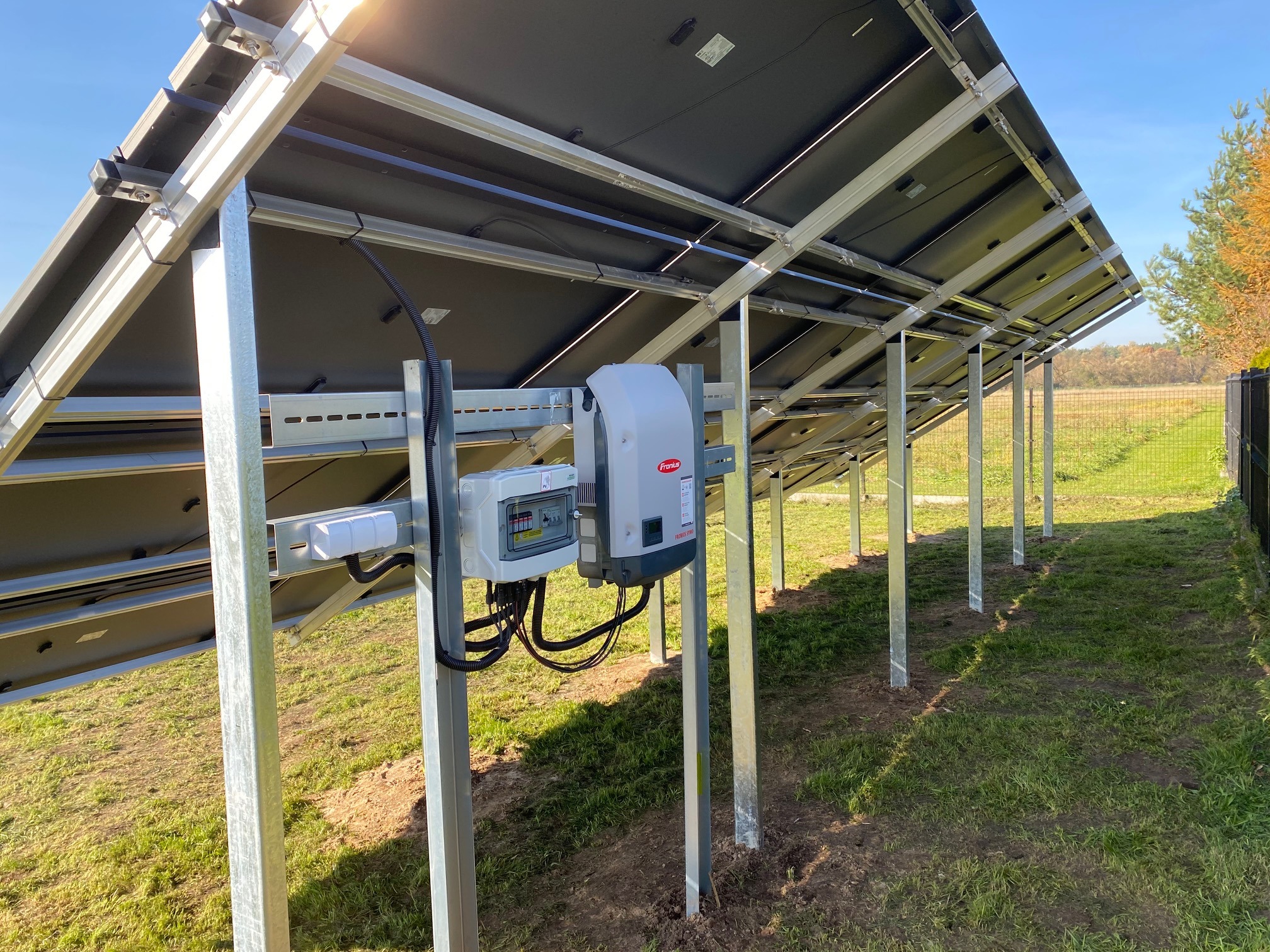 Widok od spodu na konstrukcję paneli słonecznych z zamontowanym inwerterem Fronius i skrzynką przyłączeniową na metalowych wspornikach, w tle trawa i ogrodzenie.