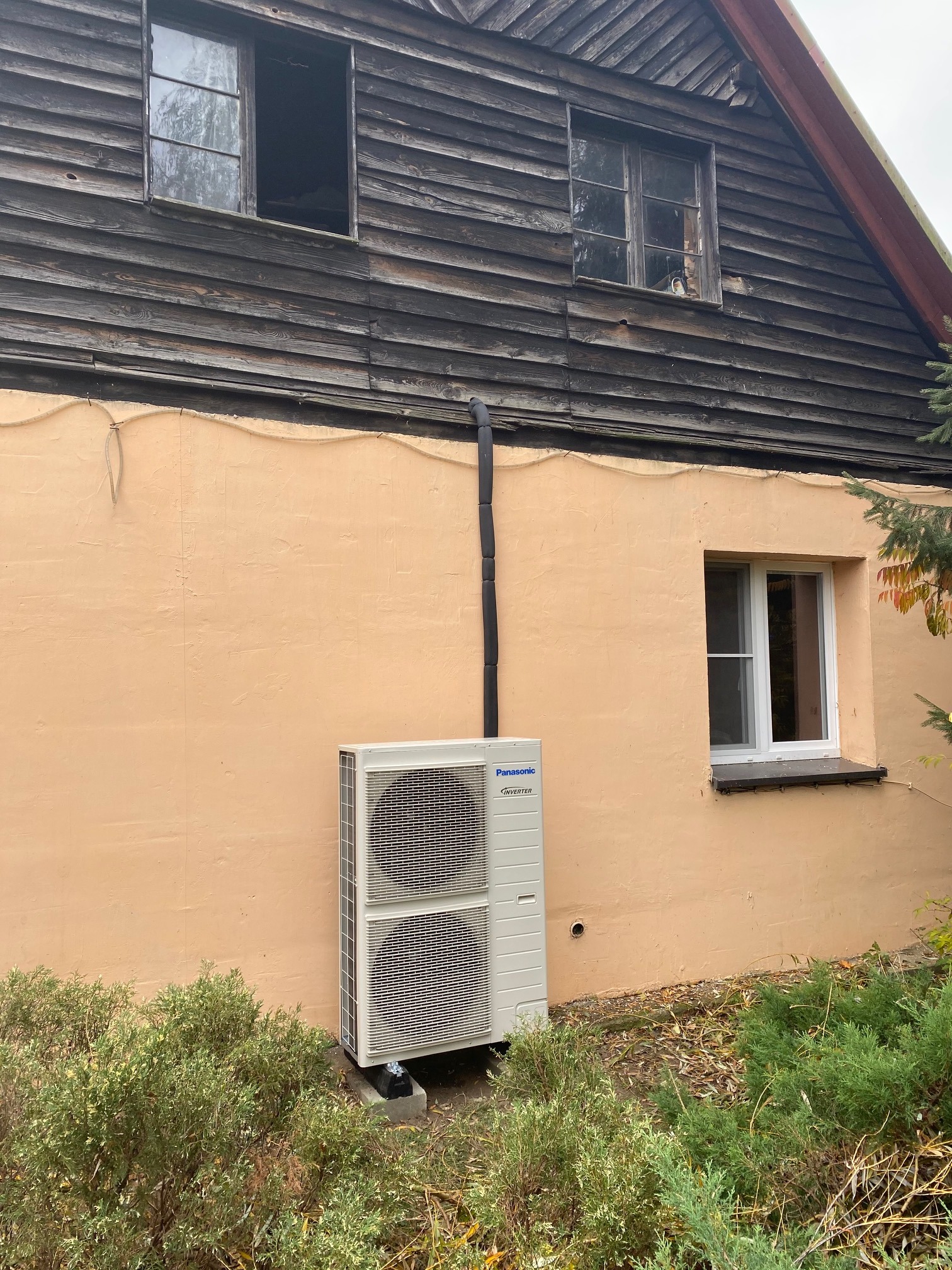 Biała jednostka zewnętrzna z czarną rurą na tle beżowej ściany budynku z drewnianą nadbudówką i oknami