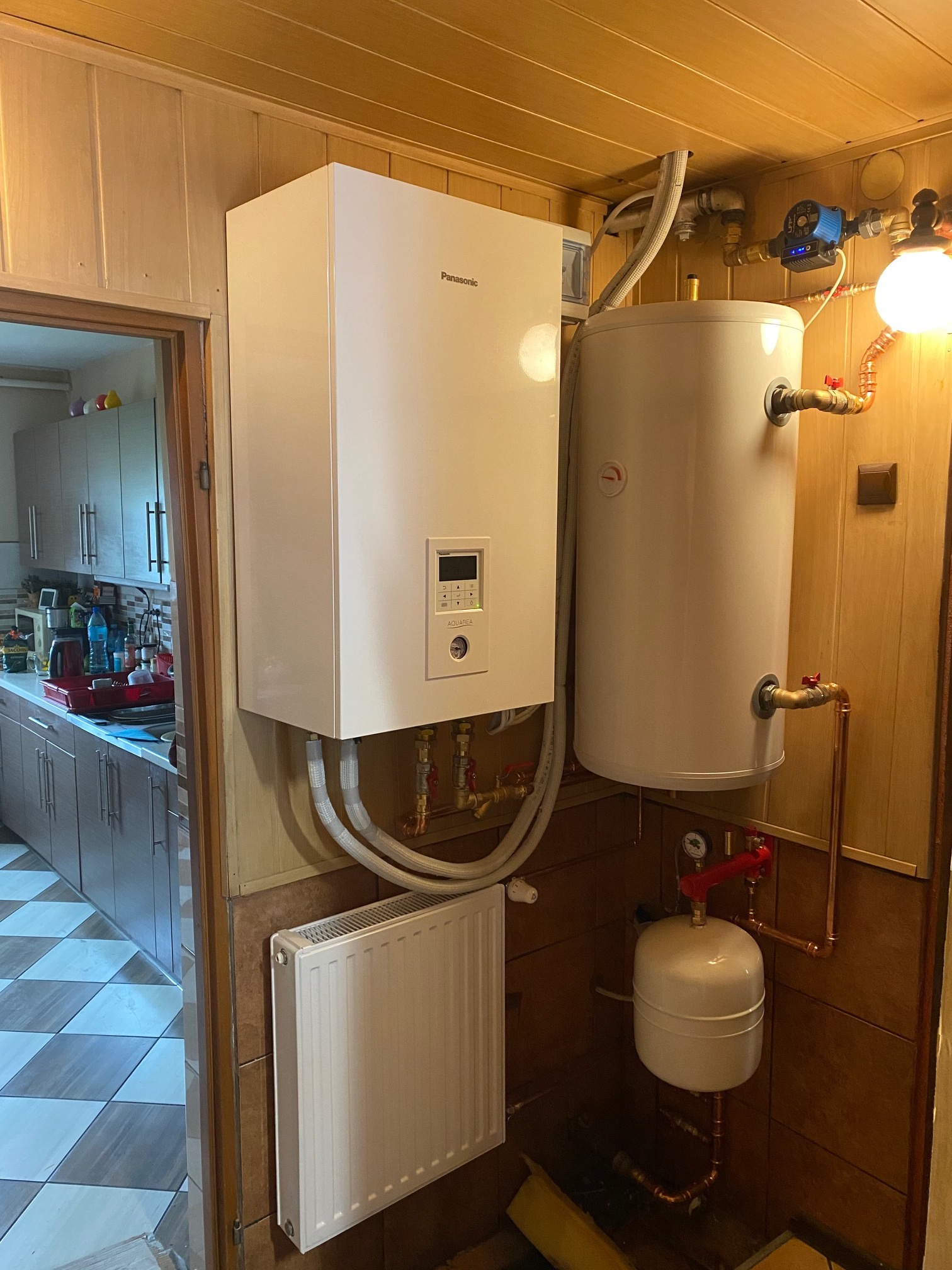 Biały, wiszący piec grzewczy Panasonic Aquarea z podłączonym zasobnikiem ciepłej wody i grzejnikiem panelowym w pomieszczeniu wykończonym boazerią i brązowymi płytkami.