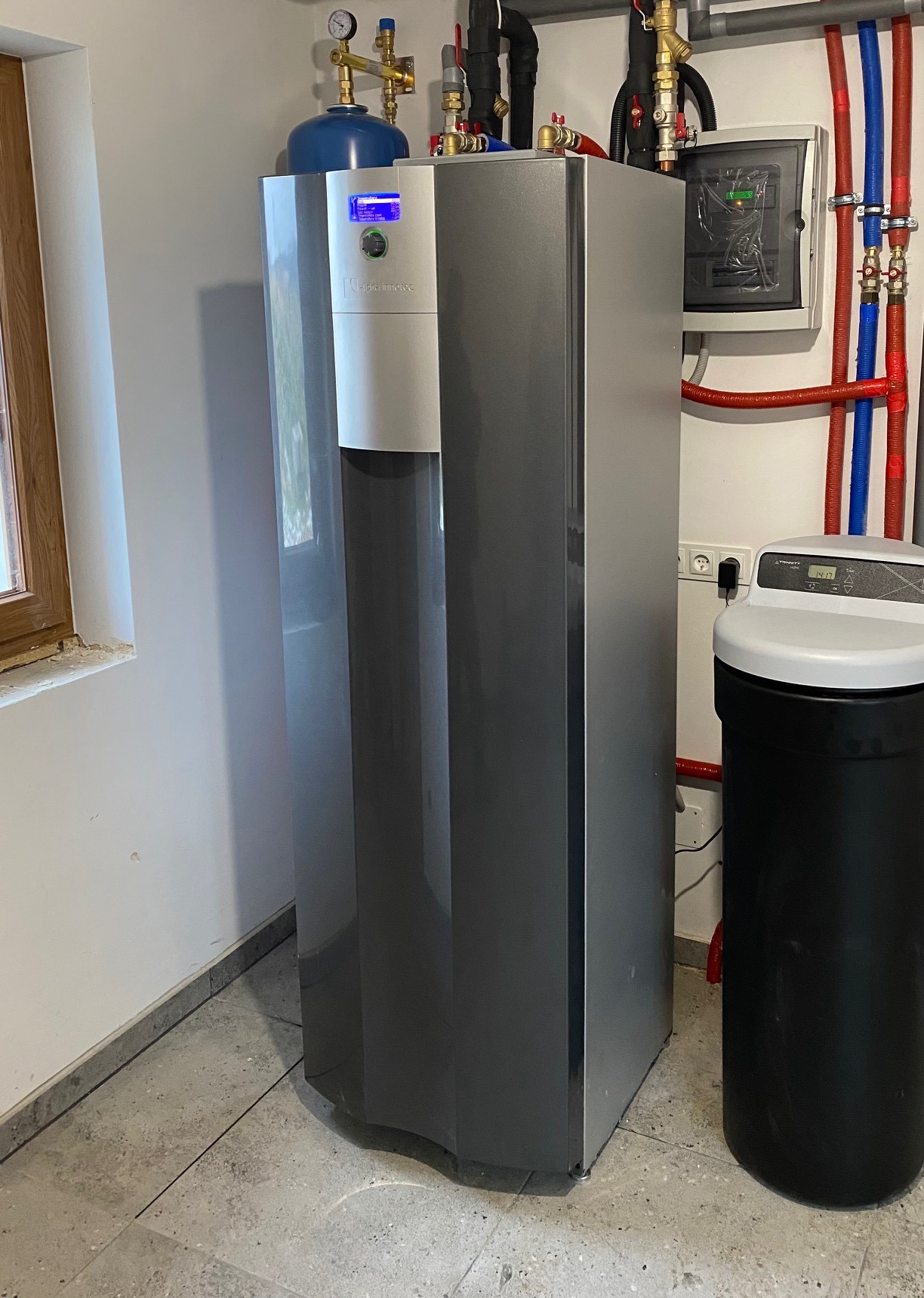 Szara jednostka wewnętrzna z niebieskim zbiornikiem ciśnieniowym oraz czarnym zmiękczaczem wody w kotłowni z instalacją hydrauliczną na ścianie.