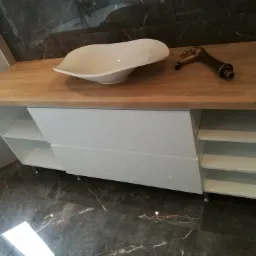 Nowoczesna łazienka z blatem z jasnego drewna, asymetryczną białą umywalką nablatową i mosiężną baterią w stylu retro, na tle płytek imitujących ciemny marmur.