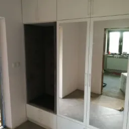 Biała szafa z lustrami i otwartą wnęką z szarą tapicerowaną półką, widoczna na tle remontowanego pomieszczenia.